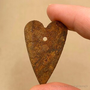 Puede incluir: Un colgante de metal en forma de coraz&oacute;n con un acabado marr&oacute;n oxidado. El colgante tiene un peque&ntilde;o agujero en la parte superior para colgarlo. La URL de la tienda de Etsy es visible en la parte inferior de la imagen.