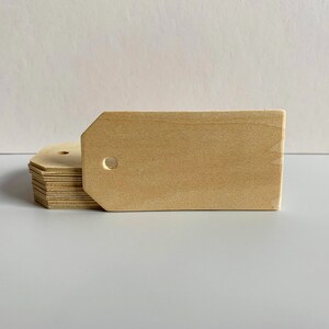 May include: A stack of unfinished wooden tags with a hole in the top. The tags are rectangular with rounded corners and are made of light brown wood.
