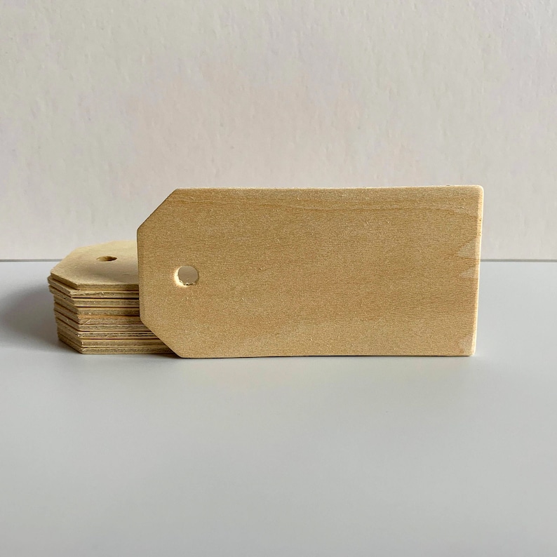 May include: A stack of unfinished wooden tags with a hole in the top. The tags are rectangular with rounded corners and are made of light brown wood.