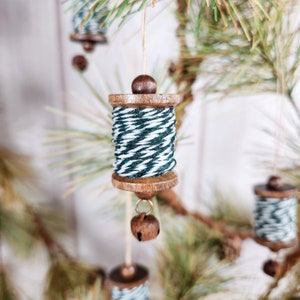 May include: A set of wooden spool ornaments with green and white striped twine, each with a small bell hanging from the bottom. The spools are hanging from a pine tree branch.