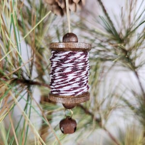 May include: A wooden spool ornament with a red and white striped twine, hanging from a pine tree branch with a small brown bell.