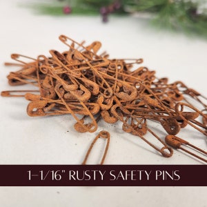May include: A pile of rusty safety pins, each measuring 1-1/16 inches. The pins are a warm, reddish-brown color, with visible signs of oxidation. The image is a close-up, showing the texture and detail of the aged metal. The text at the bottom reads "1-1/16" RUSTY SAFETY PINS."