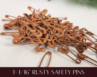 May include: A pile of rusty safety pins, each measuring 1-1/16 inches. The pins are a warm, reddish-brown color, with visible signs of oxidation. The image is a close-up, showing the texture and detail of the aged metal. The text at the bottom reads "1-1/16" RUSTY SAFETY PINS."