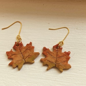 May include: A pair of gold dangle earrings featuring a brown and gold maple leaf design.