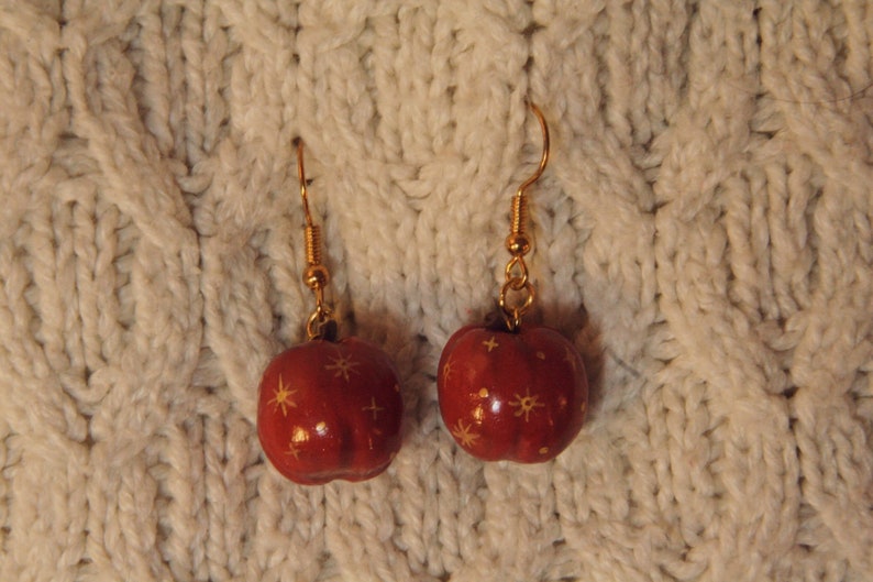 May include: A pair of red apple-shaped earrings with gold-colored hooks and starburst designs. The earrings are hanging against a textured, off-white knitted background.
