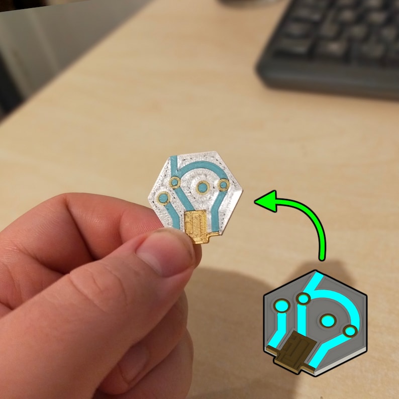 Puede incluir: Una placa de circuito hexagonal con un dise&ntilde;o azul claro y dorado. La placa tiene un acabado plateado y se sostiene en una mano.