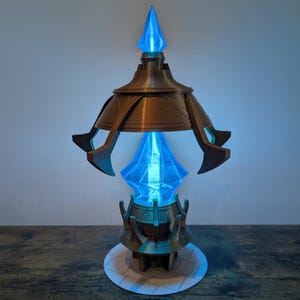 May include: A bronze-coloured, futuristic-looking lamp with a blue, glowing crystal at its centre. The lamp has a tiered design with a pointed top and a circular base. The blue light emanates from the crystal, creating a striking visual effect.