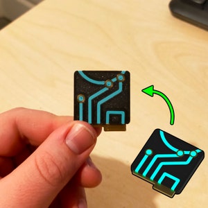 Puede incluir: Una peque&ntilde;a placa de circuito negro con un dise&ntilde;o azul y dorado. La placa tiene una superficie texturizada y es aproximadamente del tama&ntilde;o de una moneda de 20 c&eacute;ntimos de euro.