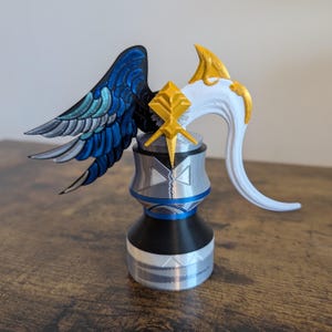 May include: A 3D printed figurine with a white and gold head, a blue and silver body, and a blue and silver wing. The figurine is standing on a wooden surface.