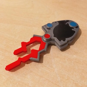 Op de afbeelding: Een 3D-geprint, abstract object met een rood, grijs, zwart en blauw kleurenschema. Het ontwerp heeft een rode, klauwachtige vorm die zich uitstrekt vanuit een grijze behuizing met een zwart midden en blauwe en rode cirkelvormige accenten.