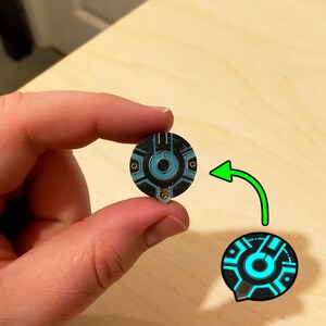 Puede incluir: Un pin circular negro y azul con un dise&ntilde;o estilizado que presenta un c&iacute;rculo con un c&iacute;rculo m&aacute;s peque&ntilde;o en su interior. El pin tiene un acento plateado.