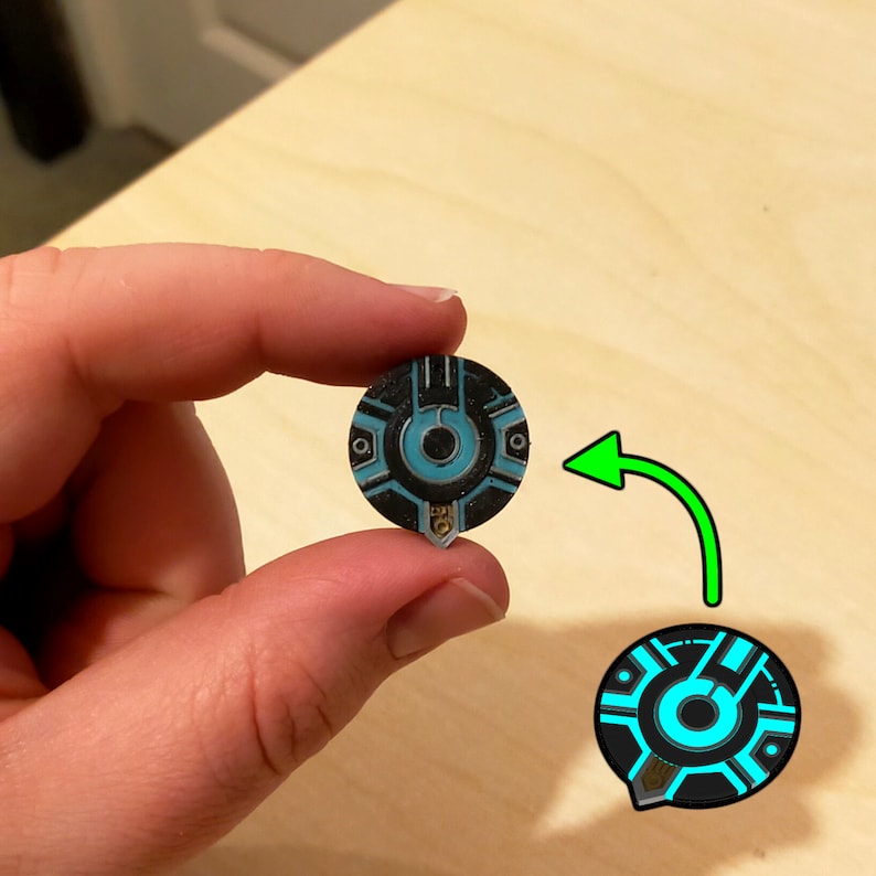 Puede incluir: Un pin circular negro y azul con un dise&ntilde;o estilizado que presenta un c&iacute;rculo con un c&iacute;rculo m&aacute;s peque&ntilde;o en su interior. El pin tiene un acento plateado.