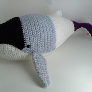 May include: A crocheted whale plush toy with a black, gray, white, and purple color scheme. The whale has a large, black eye and a long, purple tail.