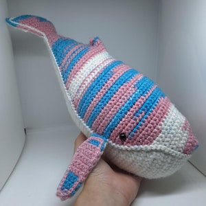 May include: A crocheted whale toy in white, pink, and blue. The whale has a striped pattern on its back and tail, with solid pink fins. The eyes are small black beads. The toy is held in a hand.