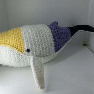May include: A crocheted whale plush toy with a yellow, white and purple colour scheme. The whale has a black tail fin and a white fin on its side.