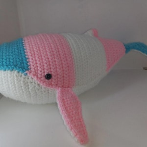Peut inclure: Une baleine en peluche fabriquée en fil avec une texture crochetée. La baleine présente des rayures horizontales bleu clair, rose et blanc. Elle possède un petit œil en perle noire sur le côté, une nageoire latérale rose et une nageoire caudale bleu clair.