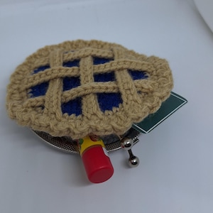 May include: A crocheted pie-shaped coin purse with a blue filling and a silver clasp. The purse is open and a red and yellow lip balm is inside.