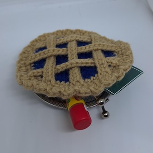 May include: A crocheted pie-shaped coin purse with a blue and tan design. The purse is open and a red tube of lip balm is inside.