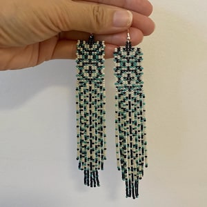 May include: A pair of long, beaded earrings with a geometric pattern in black, white, and teal. The earrings have a fringe of beads at the bottom.