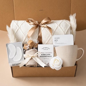 May include: A gift box containing a cream-colored textured throw blanket tied with a gold ribbon, a beige mug with vertical ridges, a box of Cartwright & Butler English Breakfast tea, a rice face mask, eye patches, caramels, and a white rose.