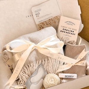 Op de afbeelding: Een geschenkdoos met een beige plaid met franjes vastgebonden met een crèmekleurig lint, een rijst-gezichtsmasker, een doos Cartwright & Butler English Breakfast-theezakjes, een paar beige geribbelde sokken, oogpatches en een witte bloemvormige kaars.