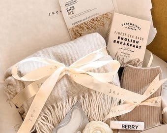 May include: A gift box containing a beige fringed throw blanket tied with a cream ribbon, a packet of Rice face mask, a box of Cartwright & Butler English Breakfast tea bags, a pair of beige ribbed socks, eye patches, and a white flower-shaped candle.