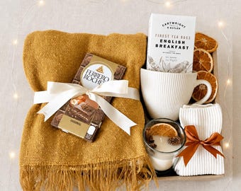 Puede incluir: Un set de regalo que incluye una manta con flecos color mostaza, una barra de chocolate Ferrero Rocher con un lazo blanco, una taza blanca acanalada, una caja de té English Breakfast de Cartwright & Butler, una vela y calcetines blancos.