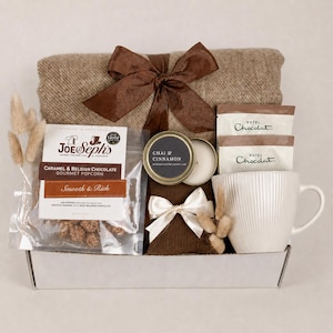 Pode incluir: Um conjunto de presente numa caixa branca contendo uma manta castanha com um laço castanho, um saco de pipocas de caramelo e chocolate belga Joe & Seph's, uma vela Chai & Cinnamon, dois pacotes Hotel Chocolat e uma caneca de cerâmica branca.