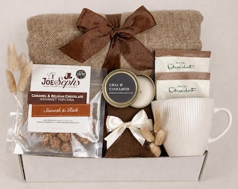 Puede incluir: Un set de regalo en una caja blanca que contiene una manta marrón con un lazo marrón, una bolsa de palomitas de caramelo y chocolate belga Joe & Seph's, una vela Chai & Cinnamon, dos paquetes de Hotel Chocolat y una taza de cerámica blanca.