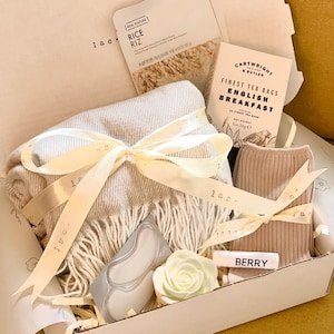 May include: A gift box filled with items for self-care and relaxation. The box includes a beige scarf with fringe, a pack of eye masks, a white rose soap, a small box of tea bags, a pair of beige socks, and a small white bar of soap with the word "BERRY" printed on it.