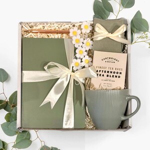 Puede incluir: Una caja de regalo llena de un diario verde, un bolígrafo, té, una taza y calcetines. El diario está atado con una cinta crema y la caja está decorada con acentos de margaritas. La caja de té dice "Finest Tea Bags Afternoon Tea Blend."
