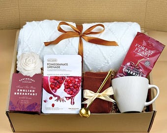 Puede incluir: Una caja de regalo llena de una toalla blanca, té, una mascarilla facial, cerezas de chocolate negro, una taza blanca y una cuchara dorada. La caja está atada con una cinta marrón.