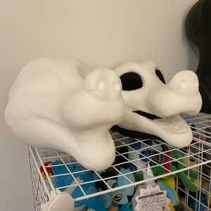 以下が含まれることがあります： 2つの白く未完成のファースーツヘッドがワイヤー棚に展示されています。1つは無垢の形で、もう1つは口と眼窩が切り抜かれています。棚にはぬいぐるみがいっぱい詰まっており、質感と色のコントラストを生み出しています。