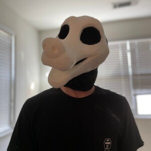 以下が含まれることがあります： 大きな黒い眼窩と突き出た鼻を持つ、白い頭蓋骨のようなスタイルのマスク。マスクは黒いTシャツの上から着用されています。マスクは小道具またはコスチュームの一部です。
