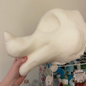 以下が含まれることがあります： コスチュームやディスプレイ用の、大きなオフホワイトの架空の生き物の彫刻された頭部。滑らかで丸みを帯びた形と顕著な眼窩があり、手で持たれています。背景には、ぬいぐるみでいっぱいのワイヤー棚があります。