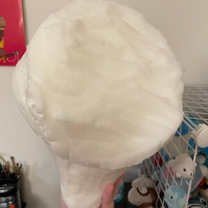 以下が含まれることがあります： 大きな白いキノコのような発泡スチロールの彫刻。彫刻は手で持たれており、丸い頭と細長い茎が特徴です。発泡スチロールは、質感のある多孔質の表面を持っています。