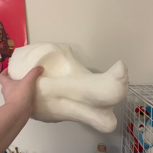 以下が含まれることがあります： 手で持たれた、オフホワイトの大きな彫刻のドラゴンヘッド。頭部には、目立つ頬骨、鼻面、滑らかな質感が特徴です。この彫刻は、コスチュームやアートプロジェクトのコンポーネントである可能性があります。