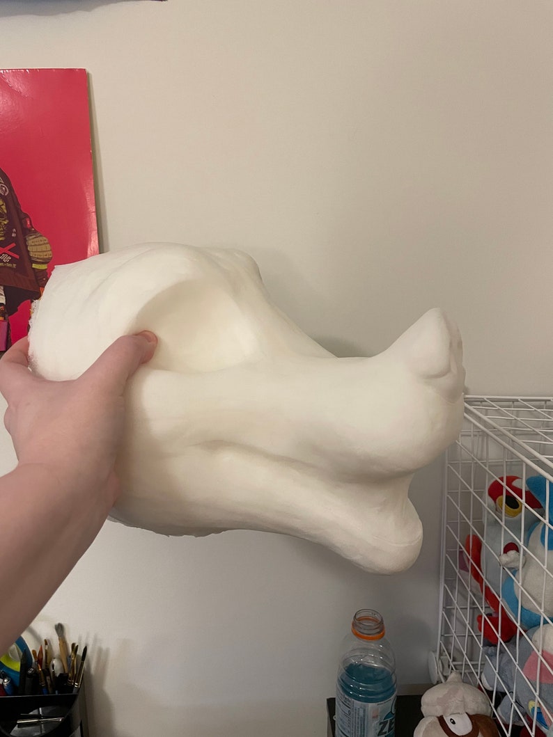 以下が含まれることがあります： 手で持たれた、オフホワイトの大きな彫刻のドラゴンヘッド。頭部には、目立つ頬骨、鼻面、滑らかな質感が特徴です。この彫刻は、コスチュームやアートプロジェクトのコンポーネントである可能性があります。