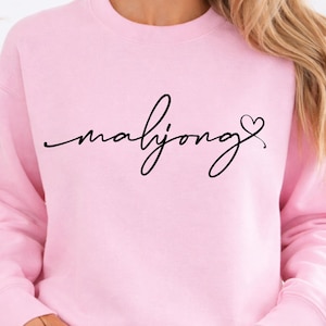 May include: A light pink sweatshirt with the word "mahjong" in a black cursive font, with a small heart symbol. The sweatshirt is a crew neck style.