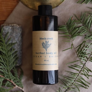 May include: A black bottle of "Black Creek Herbal Body Oil" with a label featuring a botanical illustration. The label reads "Cedar + Balsam" and "80 ml". The bottle is surrounded by green sprigs and rests on a neutral surface.