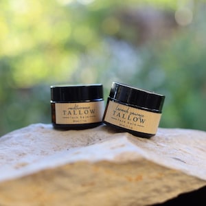 Könnte beinhalten: Zwei kleine, braune Glasbehälter mit schwarzen Deckeln, beschriftet mit "mediterranean TALLOW" und "Lavande sauvage TALLOW - face balm". Jedes Glas enthält 30 ml Produkt. Die Gläser stehen auf einer hellen, strukturierten Oberfläche.