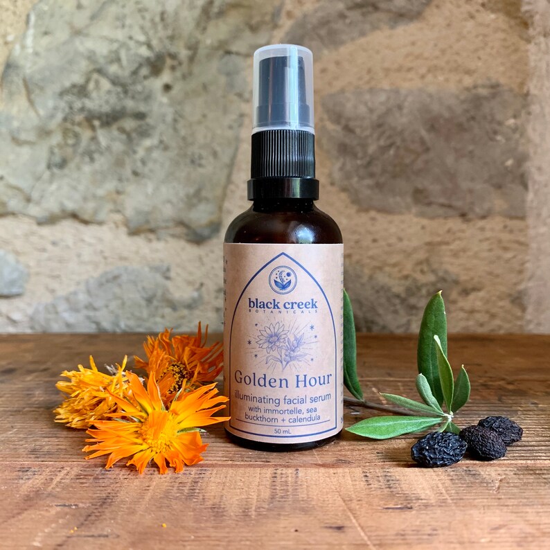 以下が含まれることがあります： 黒いポンプトップと「black creek botanicals Golden Hour illuminating facial serum with immortelle, sea buckthorn + calendula 50 ml」と書かれたラベルが付いた茶色のガラス瓶。瓶はオレンジの花と暗いベリーのある木製の表面の上に置かれています。