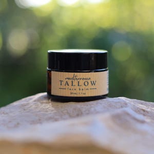 Puede incluir: Un pequeño frasco redondo de bálsamo facial con tapa negra y etiqueta marrón. La etiqueta dice "mediterranea TALLOW face balm" e indica el contenido como 30 ml / 1,1 oz. El frasco está sobre una superficie clara y texturizada.