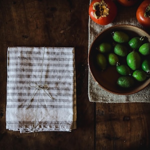 Op de afbeelding: Een stapel gevouwen, gestreepte linnen servetten, samengebonden met touw, naast een kom met groene feijoa's en verschillende oranje kaki's. De items zijn gerangschikt op een donker houten oppervlak, wat een rustieke, natuurlijke uitstraling creëert.