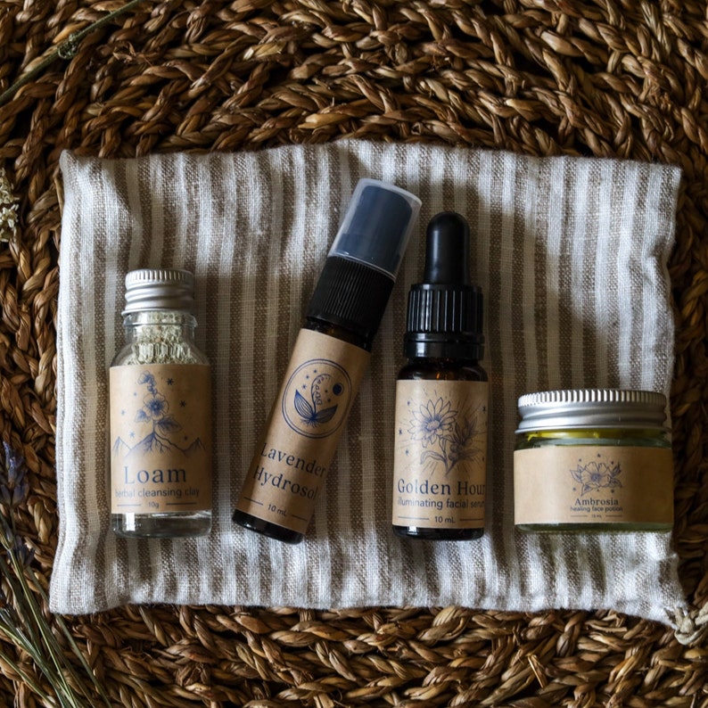 May include: Four small bottles and a jar of natural skincare products on a striped fabric surface. The bottles are labeled "Loam", "Lavender Hydrosol", "Golden Hour", and "Ambrosia".