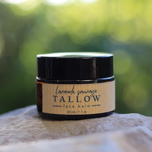 Op de afbeelding: Een kleine, ronde pot gezichtsbalsem met een zwart deksel en een bruin etiket. Op het etiket staat "Lavande sauvage TALLOW face balm" met het volume van 30 ml | 1,1 oz. De pot staat op een lichtgekleurd, getextureerd oppervlak.