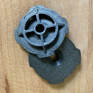 May include: Two black plastic 3D printed objects. The top object has a circular design with four spokes and a hole in the centre. The bottom object is a flat disc with a hole in the centre.