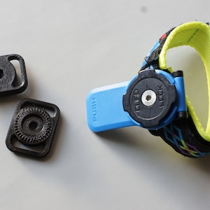 May include: A black and blue Quad Lock phone mount with a wrist strap. The mount has a blue base with the word "PUSH" and a black locking mechanism. Two additional black mounting pieces are also included.
