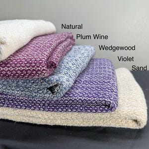 May include: A stack of five woven blankets in different colors, including natural, plum wine, wedgewood, violet, and sand.