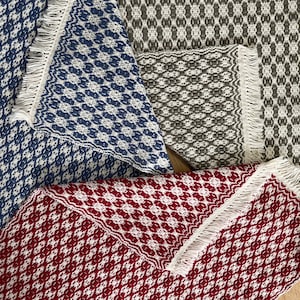 May include: Three woven placemats with fringe edges. The placemats are in shades of blue, gray, and red with a geometric pattern.